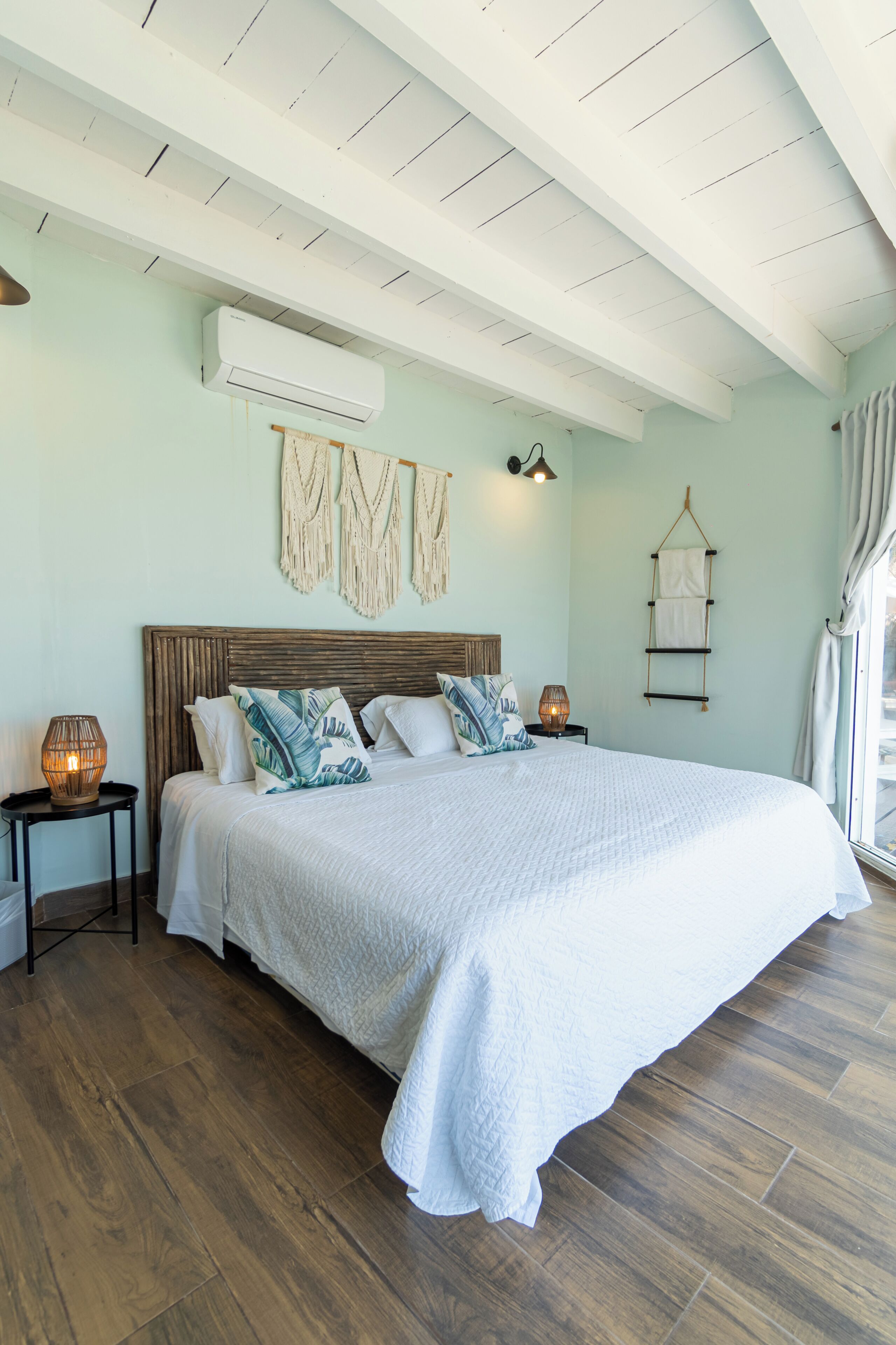 A bedroom with a large bed, wooden floor, an air conditioning unit, a wall-mounted shelf, and a bedside lamp.