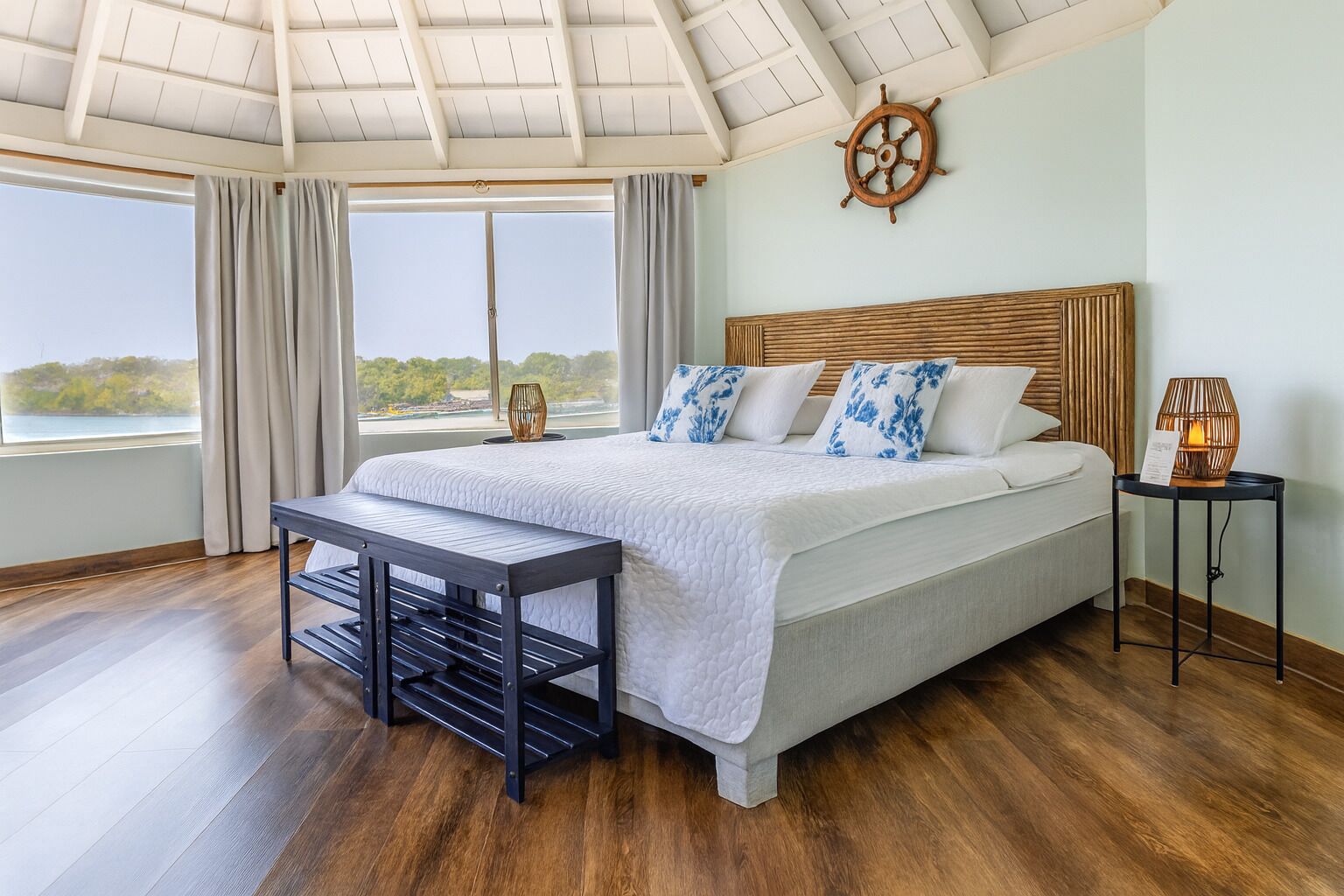 A bedroom with a large bed, a wooden headboard, a view of the outdoors, a small table, and a lamp.