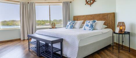A bedroom with a large bed, a wooden headboard, a view of the outdoors, a small table, and a lamp.