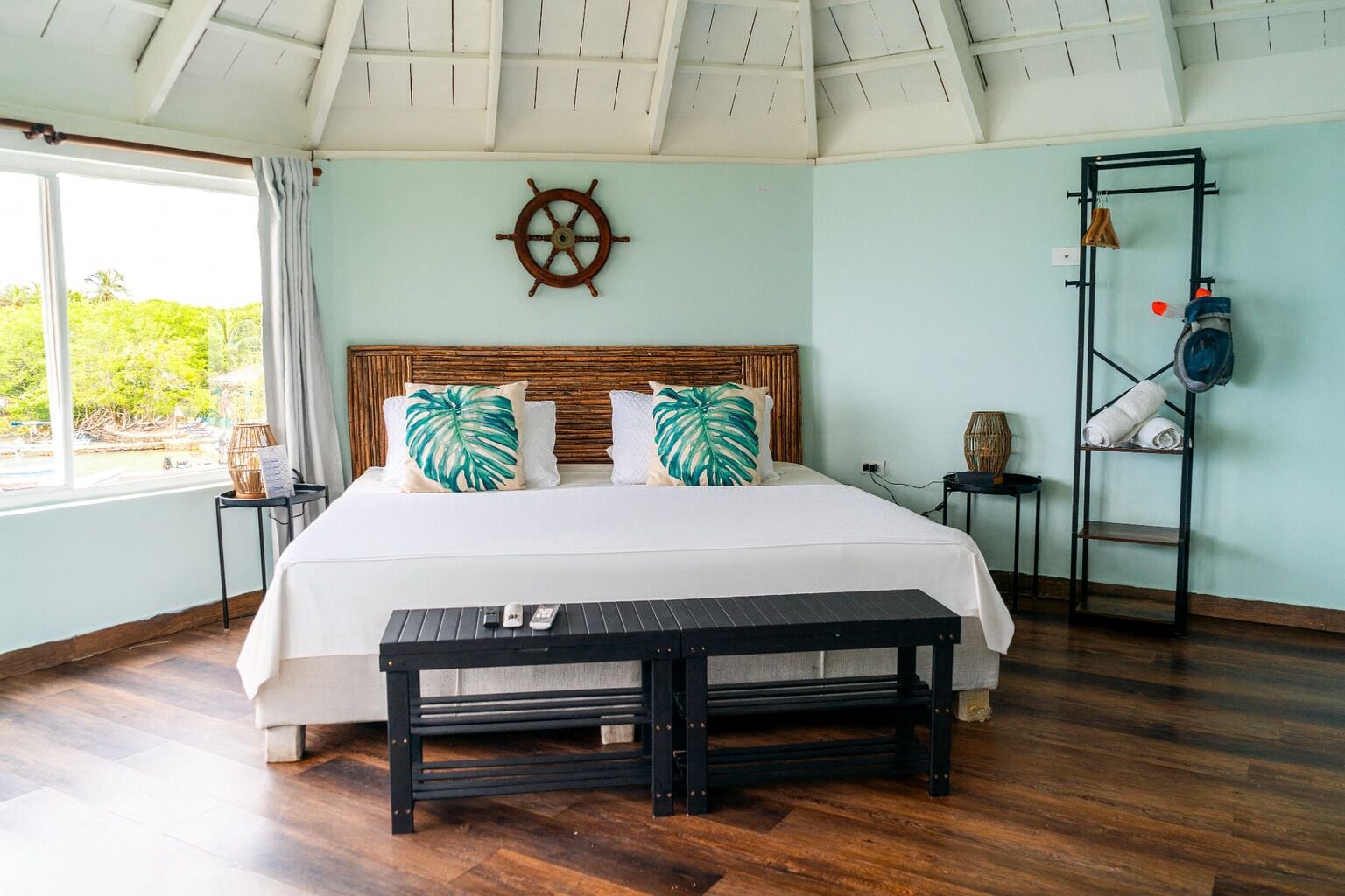 A bedroom with a large bed, a wooden headboard, a ship's wheel on the wall, a window with curtains, a small table with a vase, and a shelf with towels.