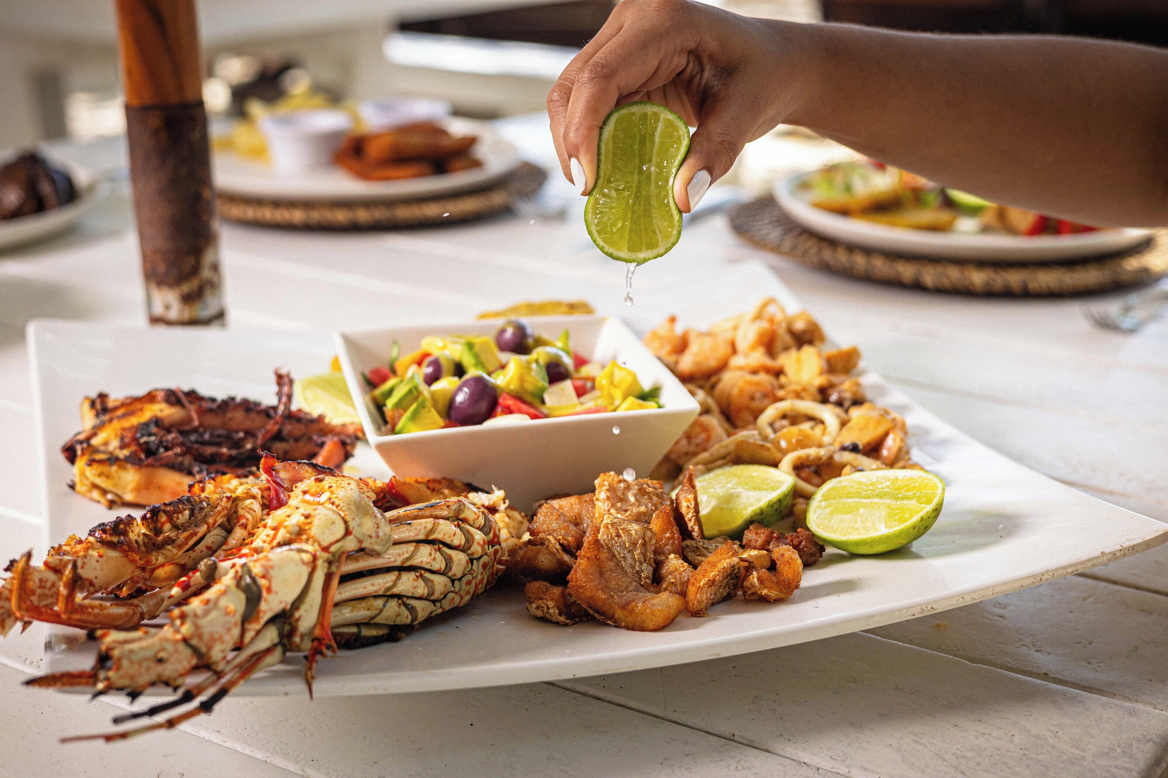 A variety of seafood dishes on a white plate, including grilled lobster and shrimp, with a hand squeezing a lime over the food.