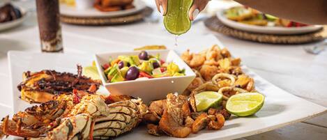 A variety of seafood dishes on a white plate, including grilled lobster and shrimp, with a hand squeezing a lime over the food.