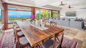 A spacious living room with a large wooden dining table, grey sofa, and ceiling fans.