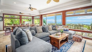 A spacious living room with a large gray sectional sofa, a wooden coffee table, and a view of the ocean through large windows.