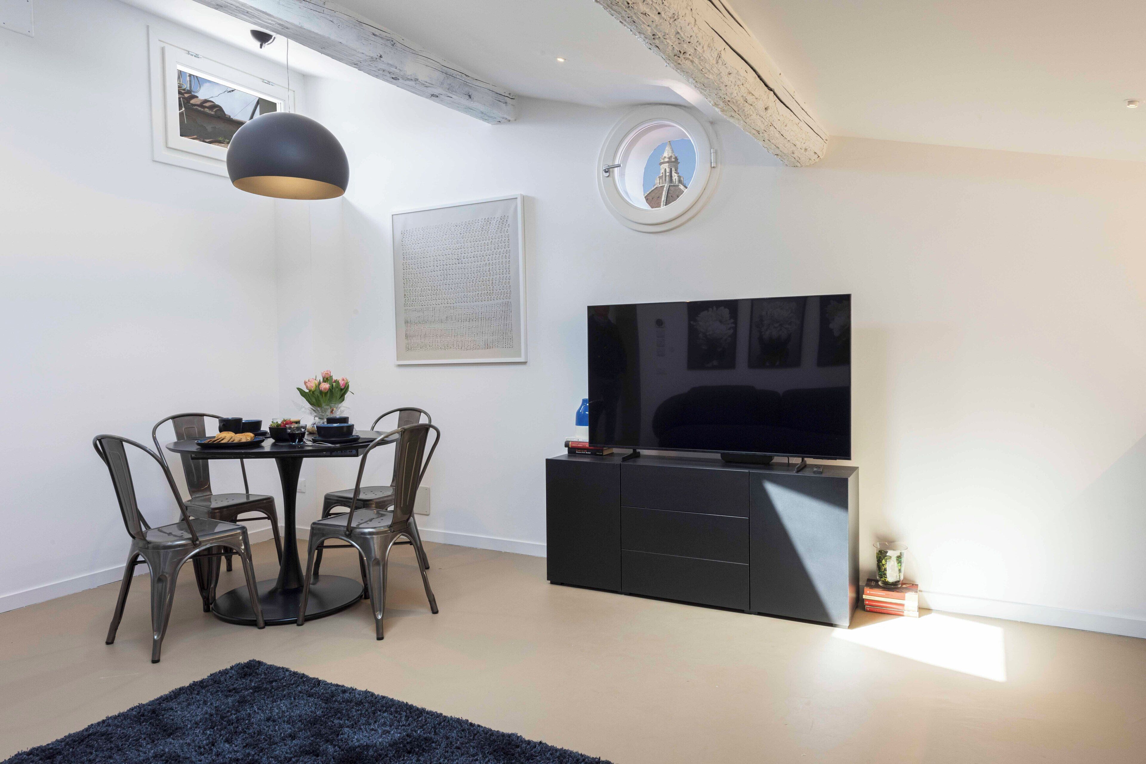 A modern living room with a dining table, chairs, a black entertainment unit, a large flat-screen TV, a round window, and a ceiling with exposed beams.