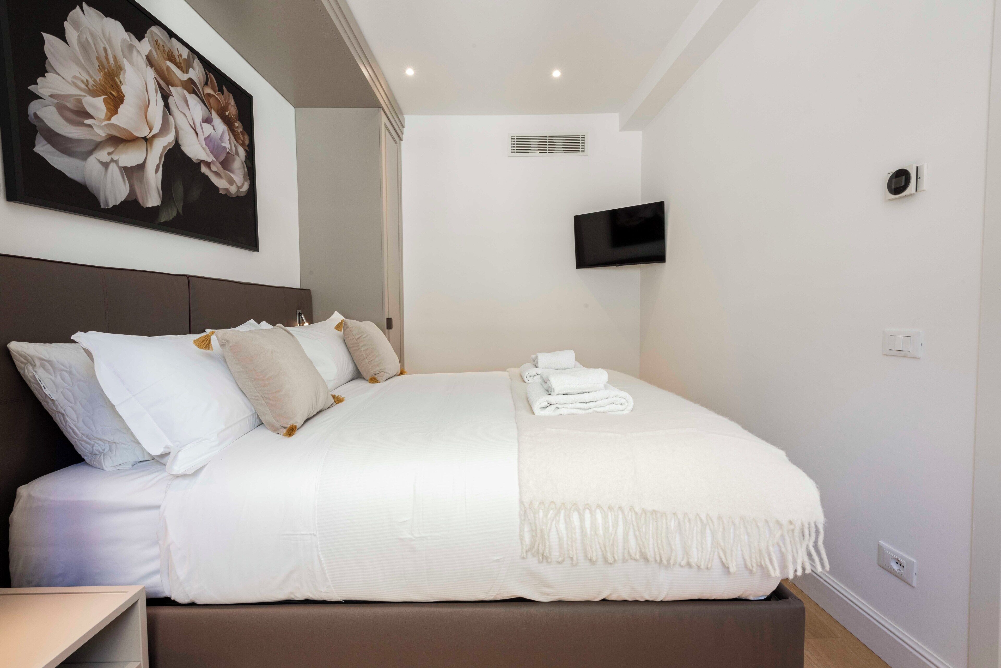 A neatly made bed with white linens and a decorative pillow, a flat-screen TV mounted on the wall, and a large floral painting above the bed.