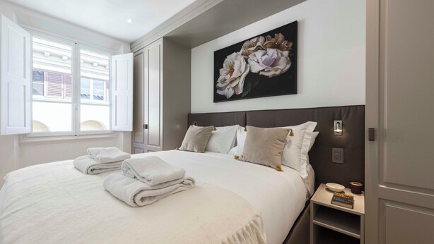 Een net opgemaakt bed met kussens en opgevouwen handdoeken, een nachtkastje met een afstandsbediening, en een aan de muur hangend bloemenstilleven.