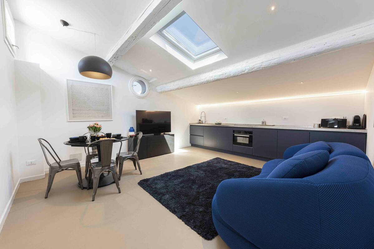 Un loft moderno con tragaluz, un comedor con sillas, un sofá azul y una cocina con armarios oscuros.