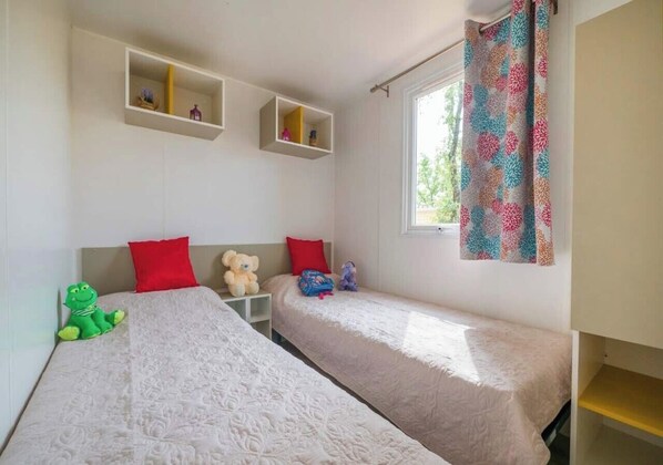 A room with two beds, each with a white bedspread, red pillows, and a small shelf above. There is a window with patterned curtains and a teddy bear on one bed. - Camping Marvilla Parks Friese Meren (Wijckel)