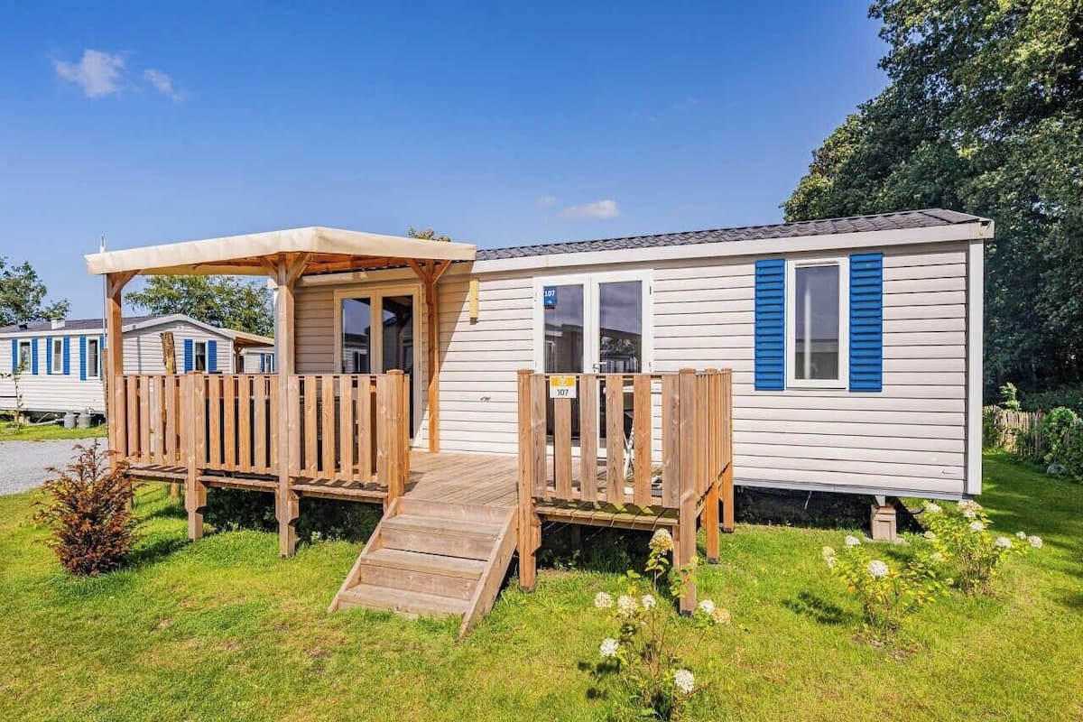 Une maison mobile dotée d’une terrasse en bois et d’une rampe d’accès, entourée d’une pelouse et d’arbres.