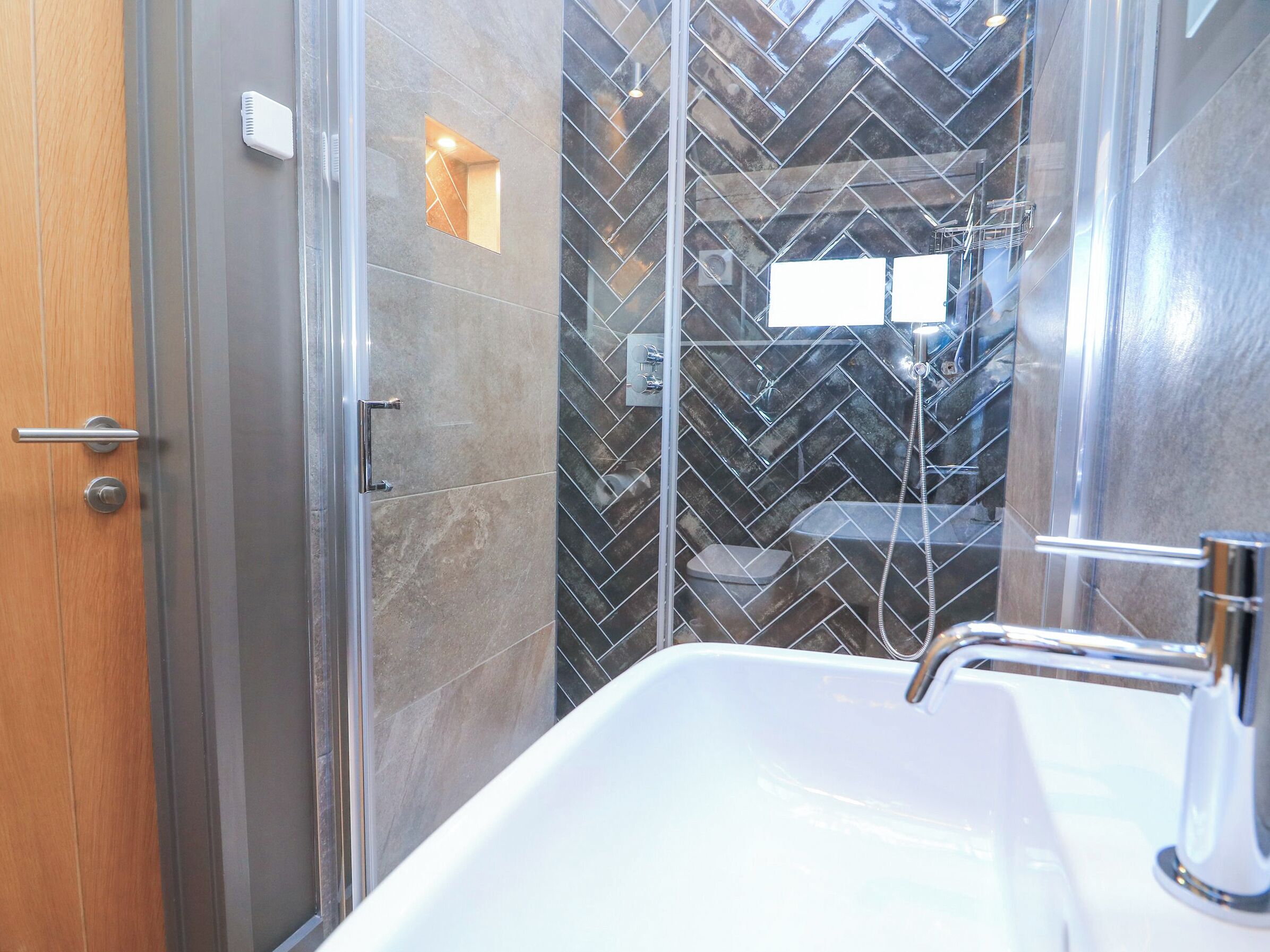 Un bagno moderno con cabina doccia in vetro, lavabo bianco e pannelli a mosaico con motivo a spina di pesce.