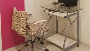 Et værelse med lyserød væg, et glasbord med computer og fladskærms-tv, en stol med leopardprint, og et blomstermotiv på væggen.