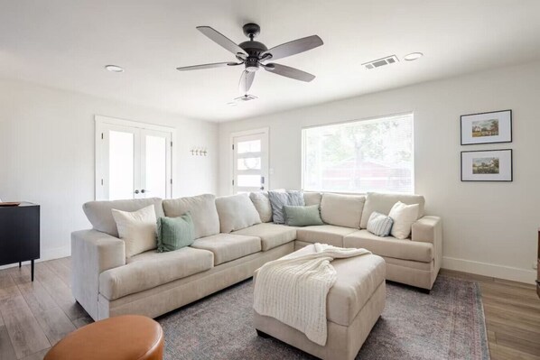 A modern living room with a sectional sofa, a ceiling fan, and large windows.