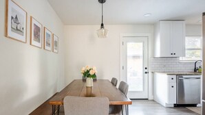 A dining area with a wooden table, chairs, and a vase of flowers. There is a kitchen with white cabinets and a stainless steel dishwasher. - Spacious 4BR Home, Central Georgetown Location! (Georgetown)