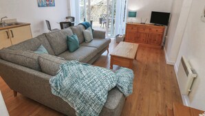 A modern living room with a sofa, a coffee table, a TV, and a kitchen area with cabinets and a sink. - Beach Court Hideaway (Holyhead)