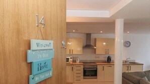 A modern kitchen with wooden cabinets, a microwave, and a sink. There is a welcome sign on the door and a clock on the wall. - Beach Court Hideaway (Holyhead)
