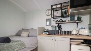 Ein kleines Schlafzimmer mit Bett, einer Kochnische, einer Uhr und einem Regal mit Küchenutensilien.