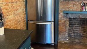 Un refrigerador de acero inoxidable en una cocina con armarios verde azulado y una pared de ladrillo.