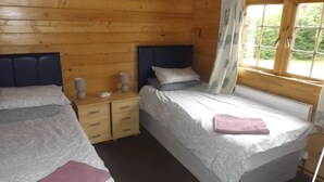 Quarto de cabana de madeira com duas camas, uma mesinha de cabeceira e uma janela com cortinas.