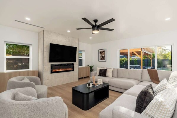 A modern living room with a fireplace, a ceiling fan, and a large window overlooking a garden.