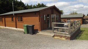 A wooden cabin with a porch, a trash bin, and a small outdoor seating area. - Spacious 3 Bedroom Cabin at Avonvale Holiday Lodge (Evesham)