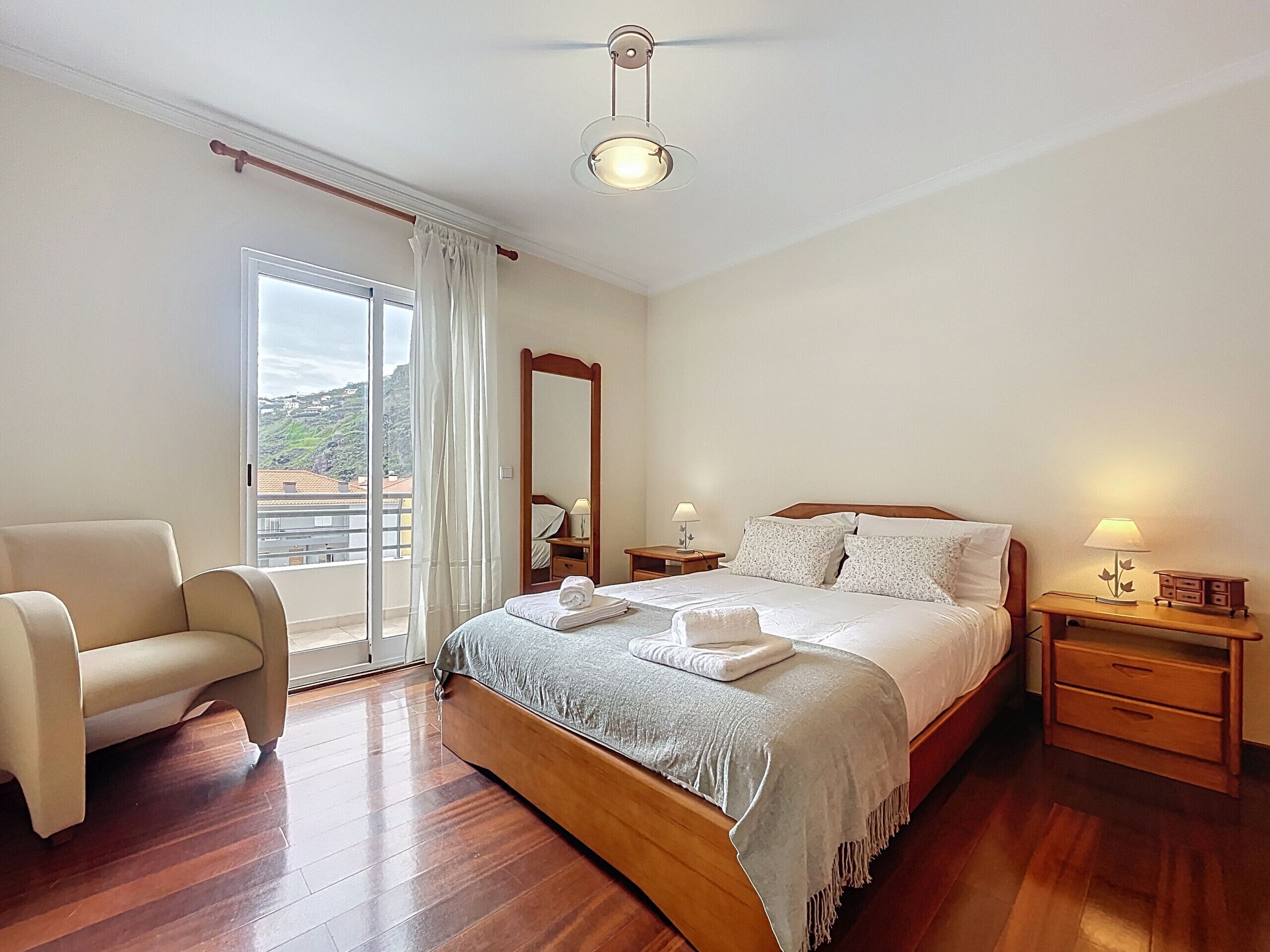 A bedroom with a bed, a chair, a nightstand, a mirror, and a window with curtains.