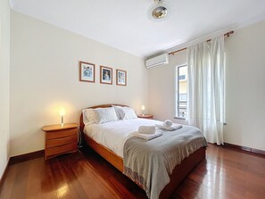 Een slaapkamer met een houten bed, twee nachtkastjes met lampen, een raam met gordijnen, en drie ingekaderde foto’s aan de muur.