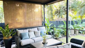 Ein überdachter Außenbereich mit einer Couch und Sesseln, umgeben von Kübelpflanzen und Blick auf einen Garten.