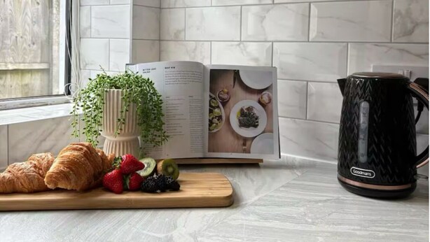 A kitchen with a marble backsplash, a wooden cutting board with croissants and fruits, a cookbook, and a black kettle.