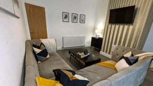 A modern living room with a grey sofa, yellow and blue pillows, a coffee table, a black side table, and a mounted TV.