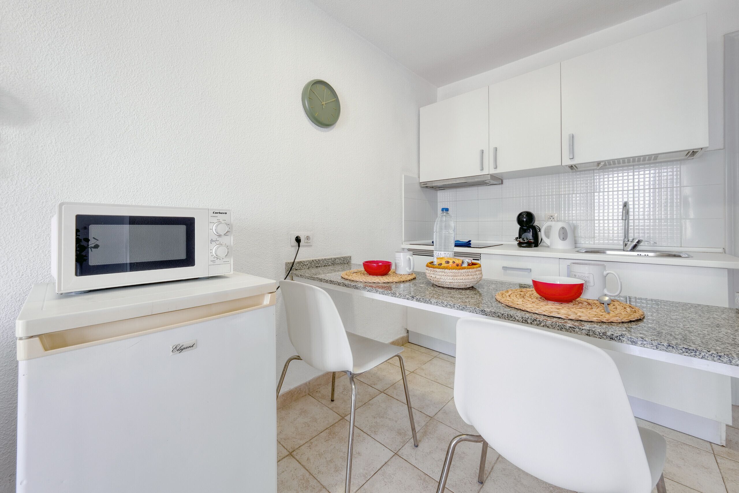 Uma cozinha compacta com armários brancos, micro-ondas, pia e um relógio na parede.