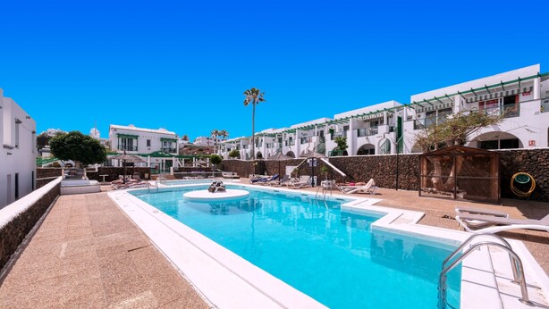 A swimming pool with lounge chairs, a stone wall, and white buildings with green accents. - Eva Vacation Home Pool & Relax - Very close to the beach (Puerto del Carmen)