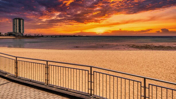 A beach with a metal railing in the foreground and a tall building in the background. - Studio el Greco next to Reducto beach (Arrecife)
