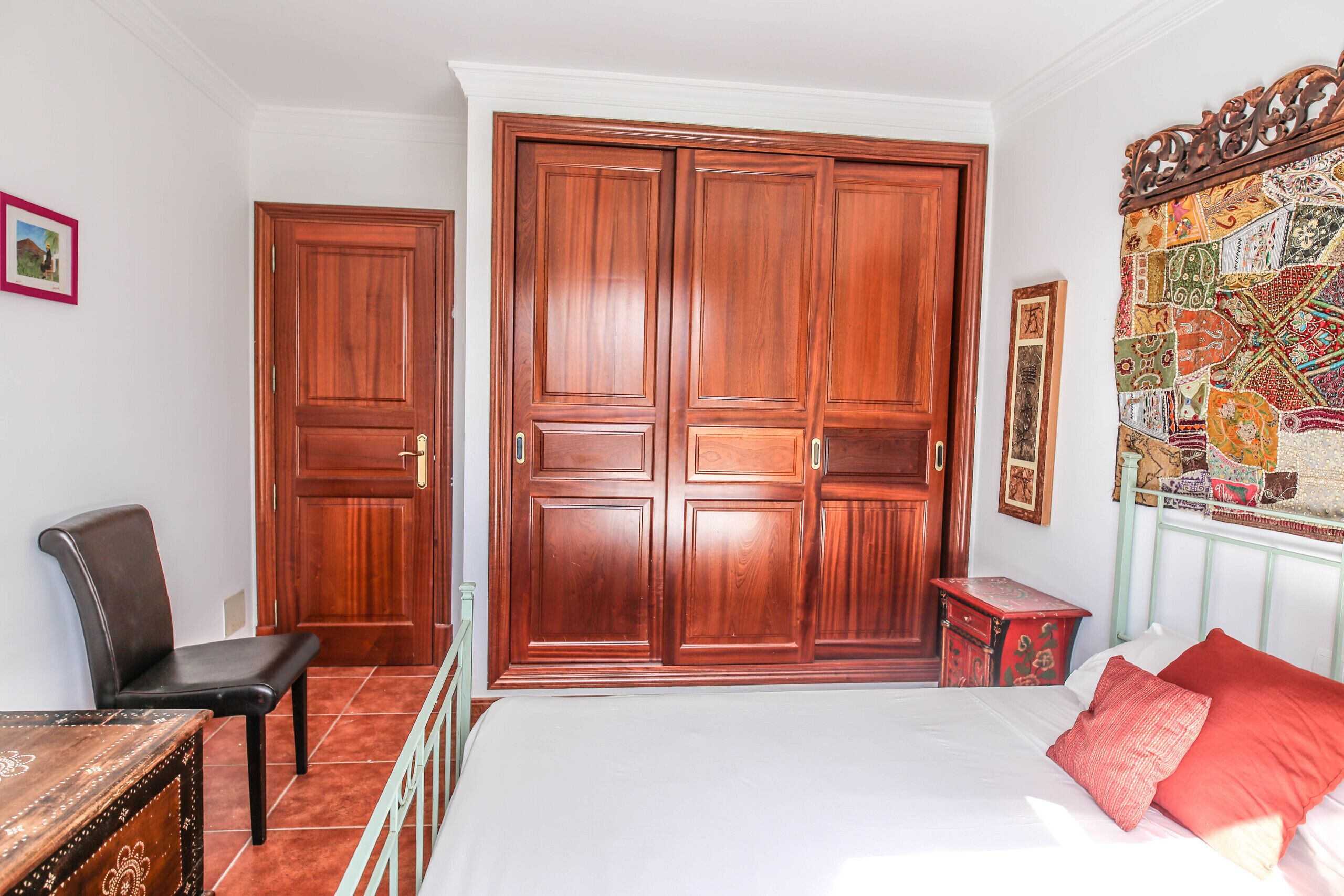 Ein Schlafzimmer mit großem Holzschrank, einem Bett mit roten Kissen, einem kleinen Nachttisch in Rot und einem Sessel.