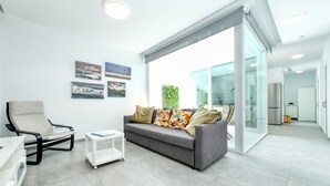 A modern living room with a grey sofa, a white armchair, and a small white side table. There are framed pictures on the wall and a glass door leading to a balcony.