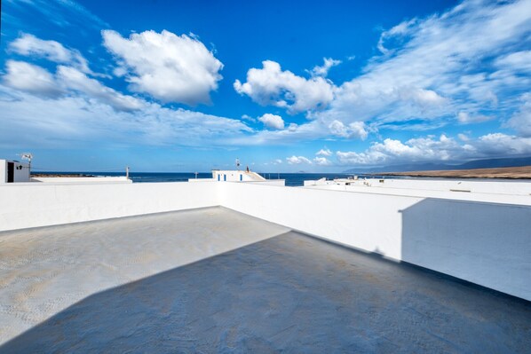 A rooftop terrace with a clear view of the sea, a white building, and a lighthouse in the distance.