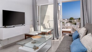 Uma sala de estar moderna com televisão de tela plana, mesa de centro de vidro, sofá e varanda com vista para a cidade.