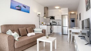 A modern living room with a brown sofa, white coffee table, and a kitchen area with white cabinets and appliances.