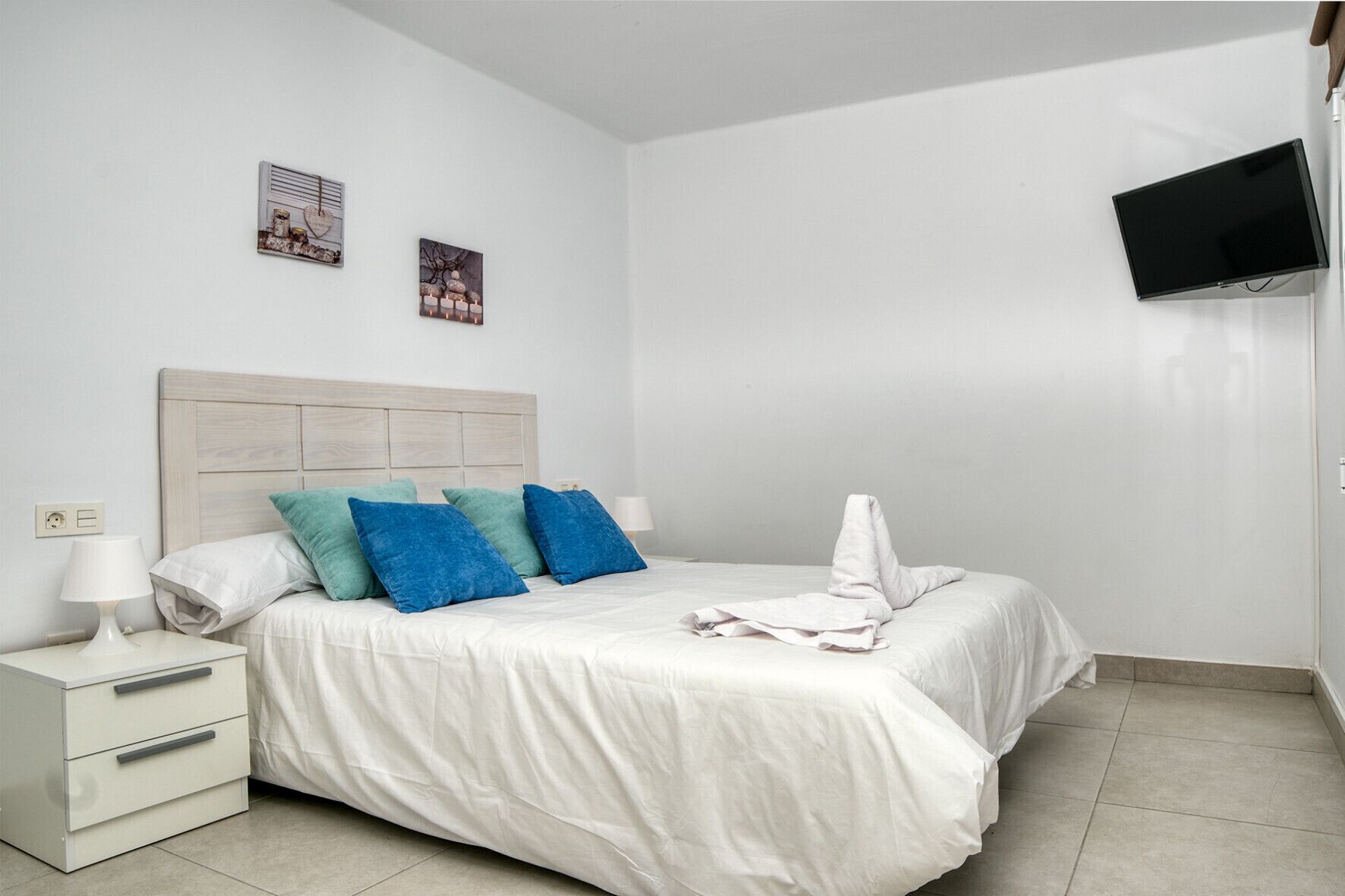 A neatly made bed with white bedding and blue pillows, a bedside table with a lamp, and a flat-screen TV mounted on the wall.