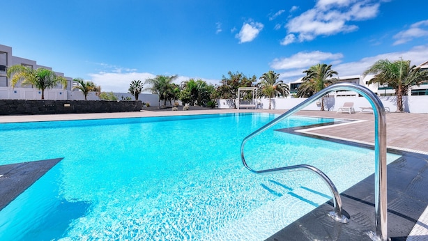 A clear blue pool with a metal ladder, surrounded by palm trees and modern buildings.
