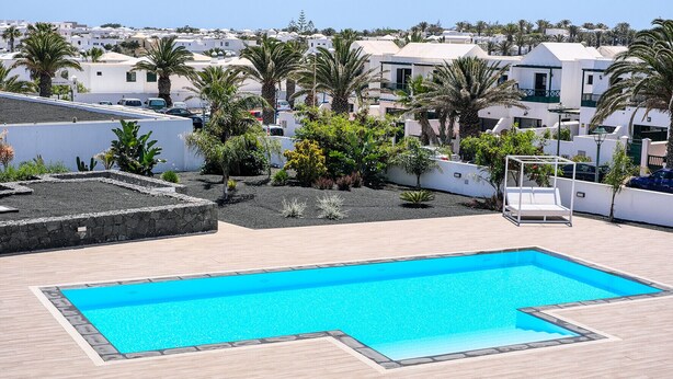 A rectangular pool with a wooden deck surrounding it, set against a backdrop of white buildings and palm trees.