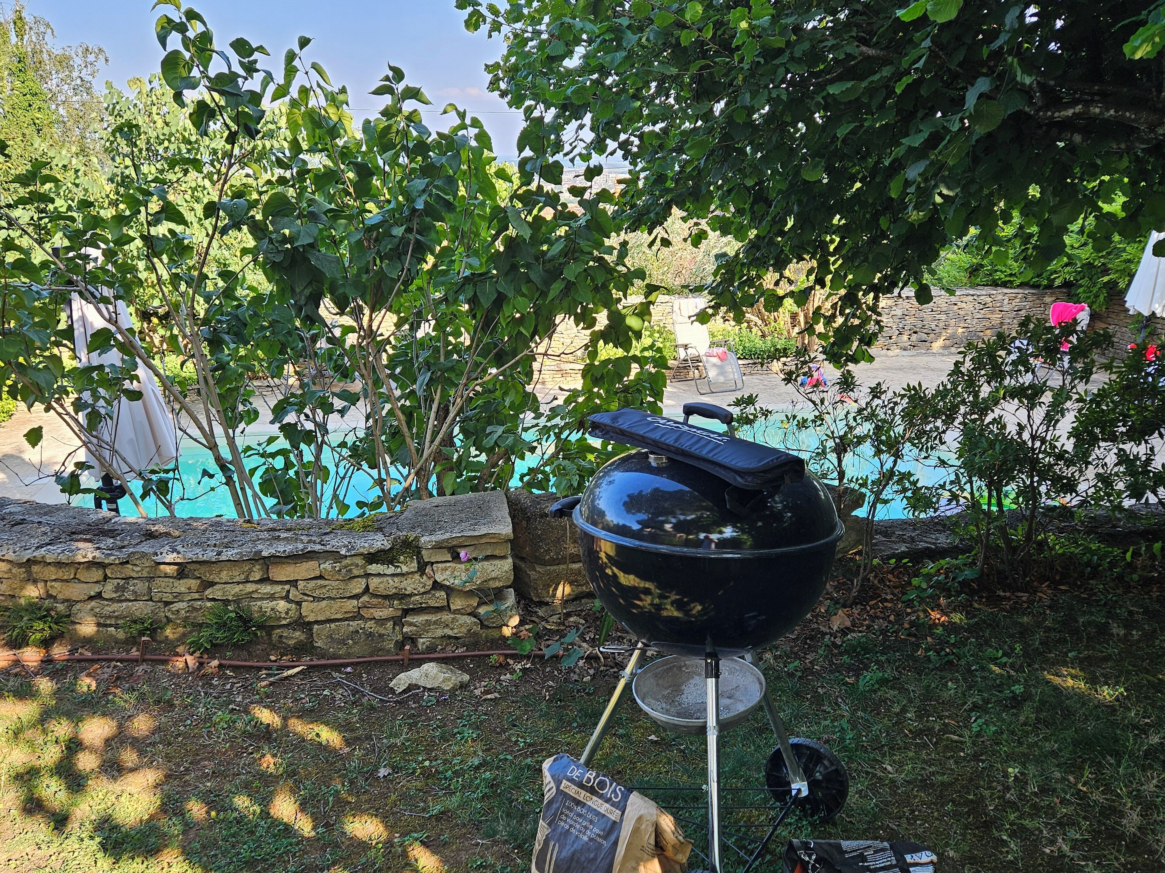 A portable grill set up in a backyard with a stone wall and a swimming pool in the background.