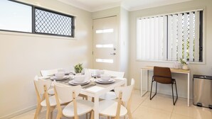 A dining area with a table set for four, white chairs, a small side table with a vase, and a window with blinds. - Smart Suites Self Check-in Apartments - 20 Darnley (Rocklea)