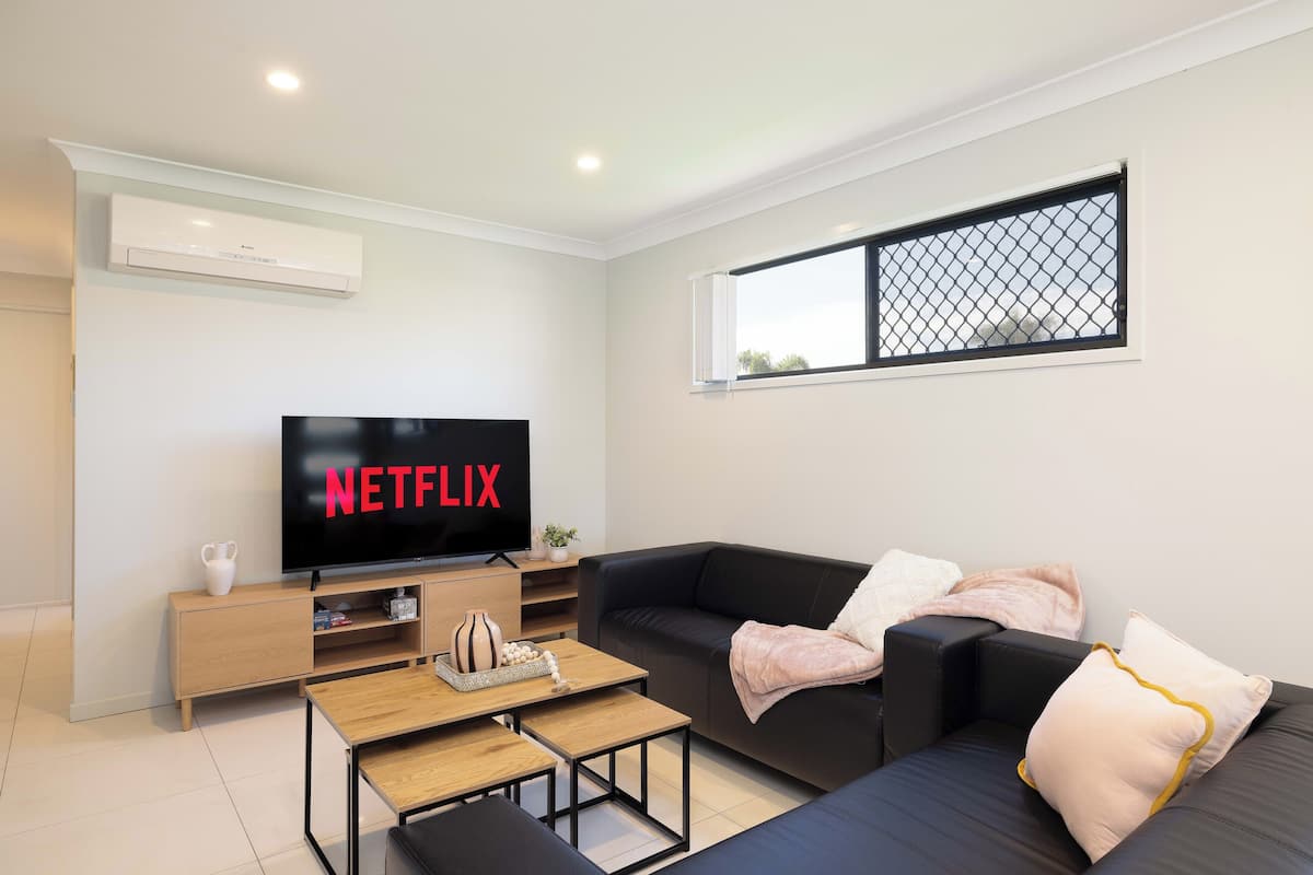 Una sala de estar moderna con un televisor de pantalla plana mostrando 'NETFLIX', un sofá negro, una mesa de centro de madera y una mesita auxiliar con adornos.