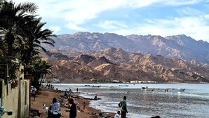 A beach with rocky mountains in the background, boats on the water, and people on the shore.