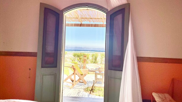 A room with a bed, a chair, and an open door leading to an outdoor area with a view of the sea and a person sitting on a chair.