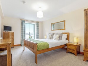 Ein Schlafzimmer mit einem Holzbett, einem Schreibtisch mit Fernseher, einem Nachttisch mit Lampe und einem Fenster mit Vorhängen.