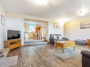 Uma sala de estar moderna com televisão de tela plana, sofá de couro, mesa de centro de madeira e uma área de refeições com cozinha ao fundo.