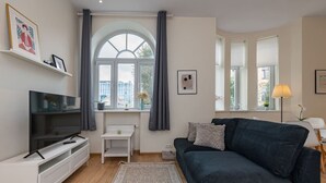 A modern living room with a flat-screen TV, a dark blue sofa, a small white table, a large window with curtains, and a floor-to-ceiling mirror.