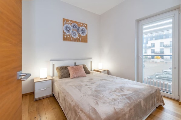 A bedroom with a bed, bedside tables, a nightstand, a lamp, a painting, and a view of the outside.